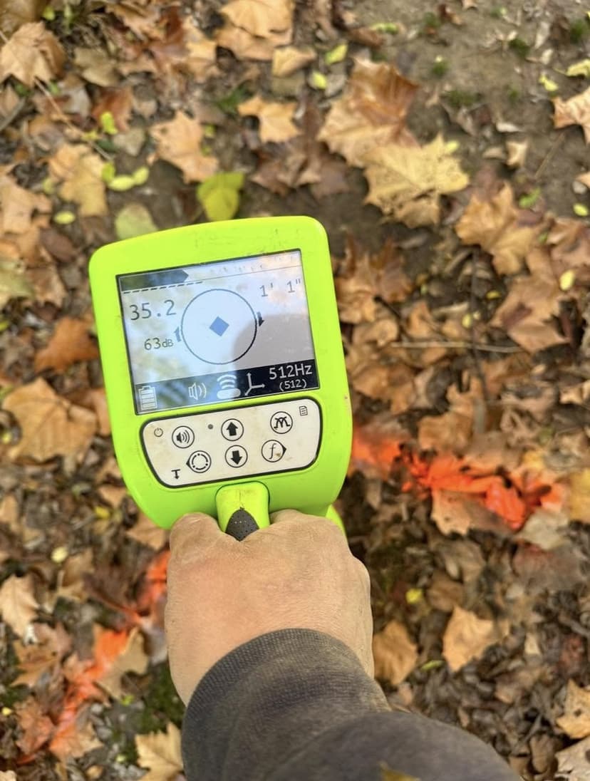 A handheld locator reads the signal from a pipe camera about a foot underground; orange paint marks the line