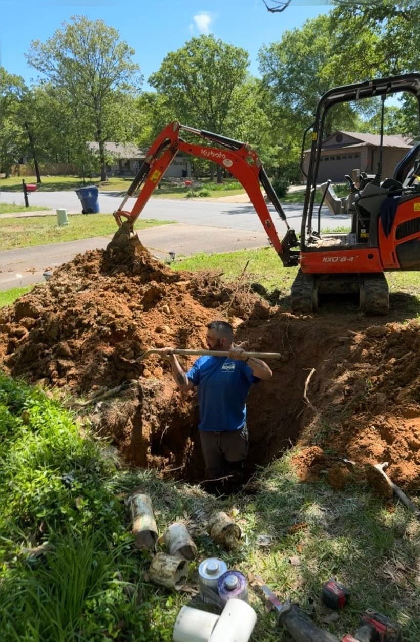 A Victory plumber digs out a buried line in a front yard, with a mini excavator behind him