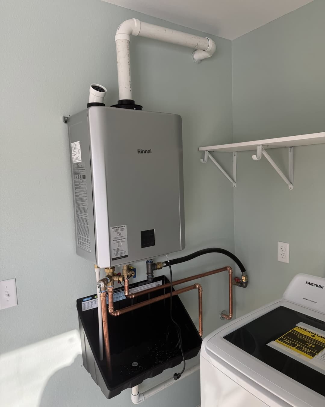 A finished Rinnai tankless water heater install in a laundry room, with copper water lines, PVC venting and a drain pan