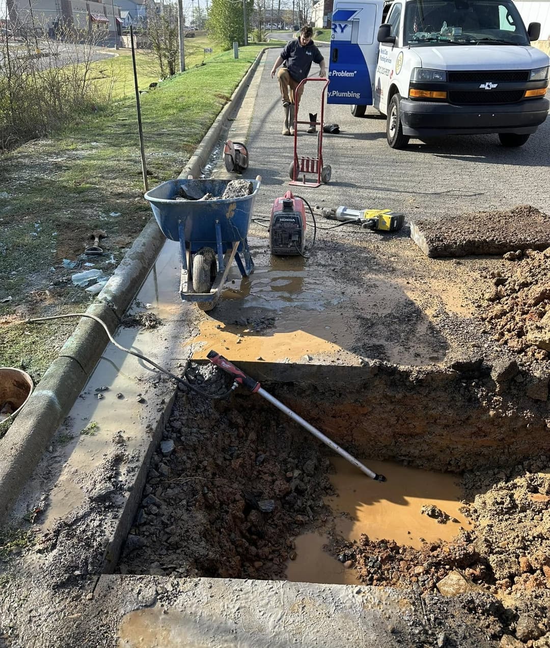 A Victory plumber in waders works a water line dig beside a curb, with the Victory van parked behind him